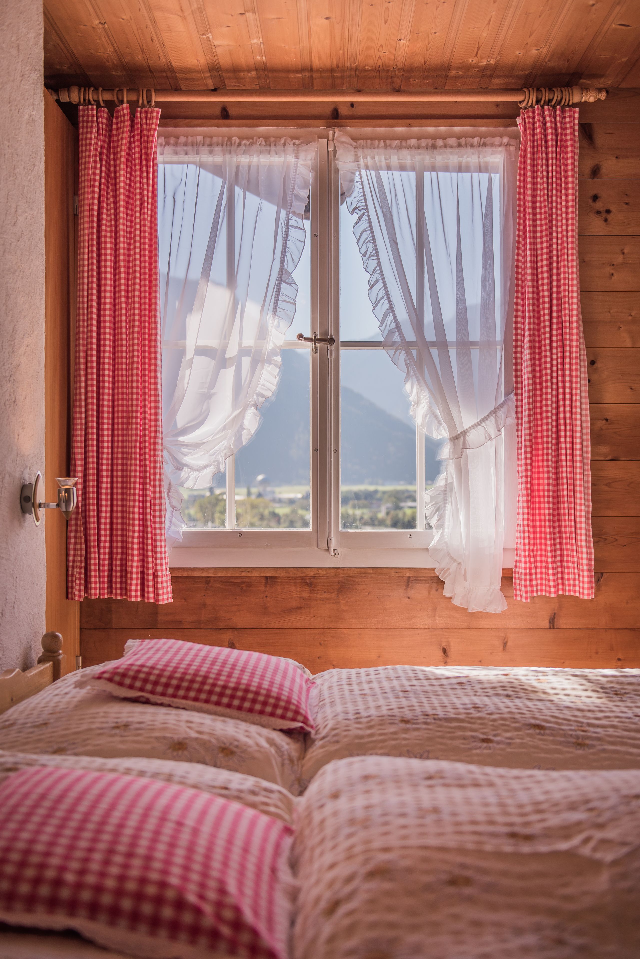 A room with a wooden ceiling, a window with red and white checkered curtains, and a bed with checkered bedding.