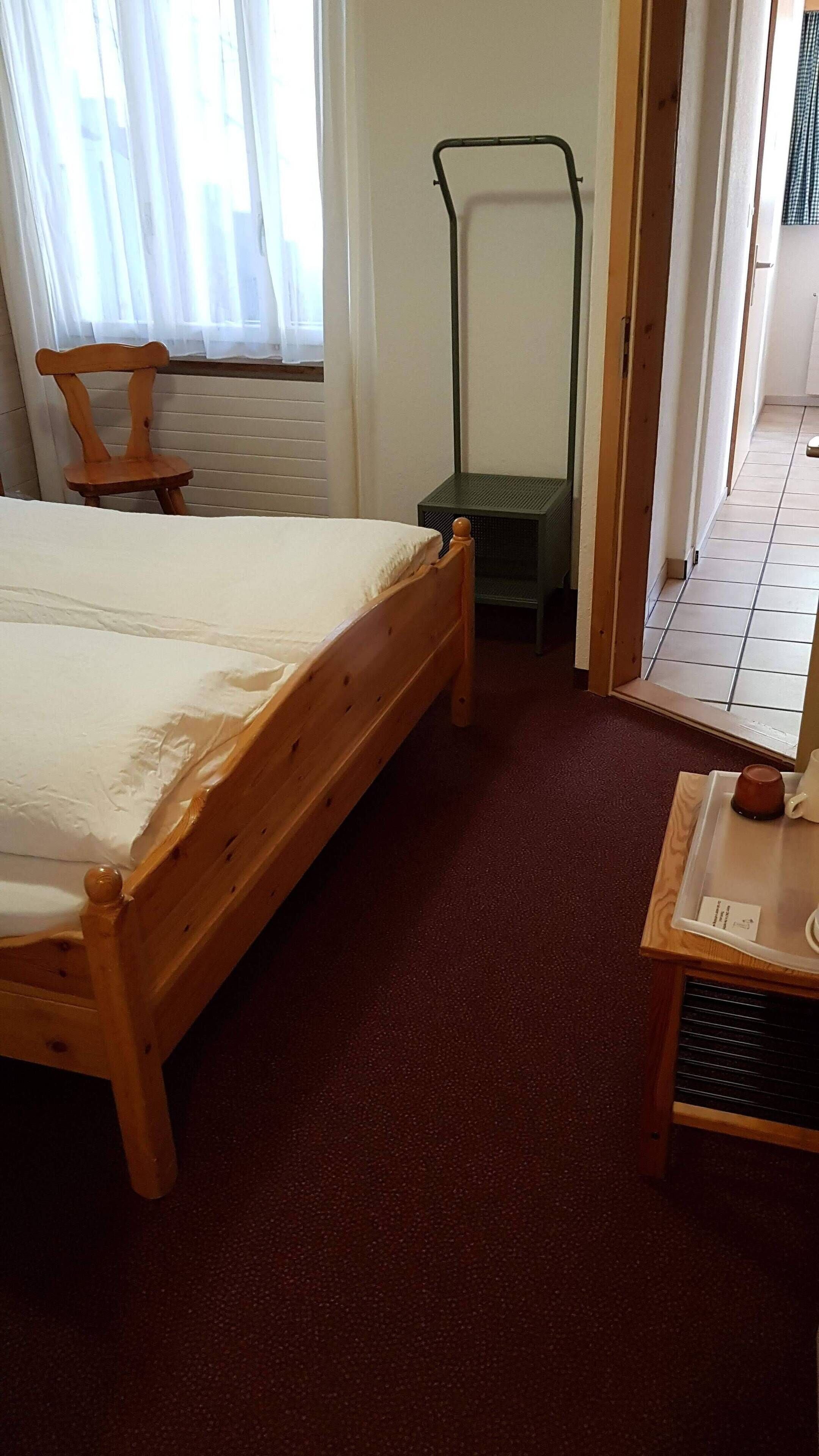 Una cama de madera en una habitación con alfombra roja, una ventana con cortinas traslúcidas y una silla de madera.