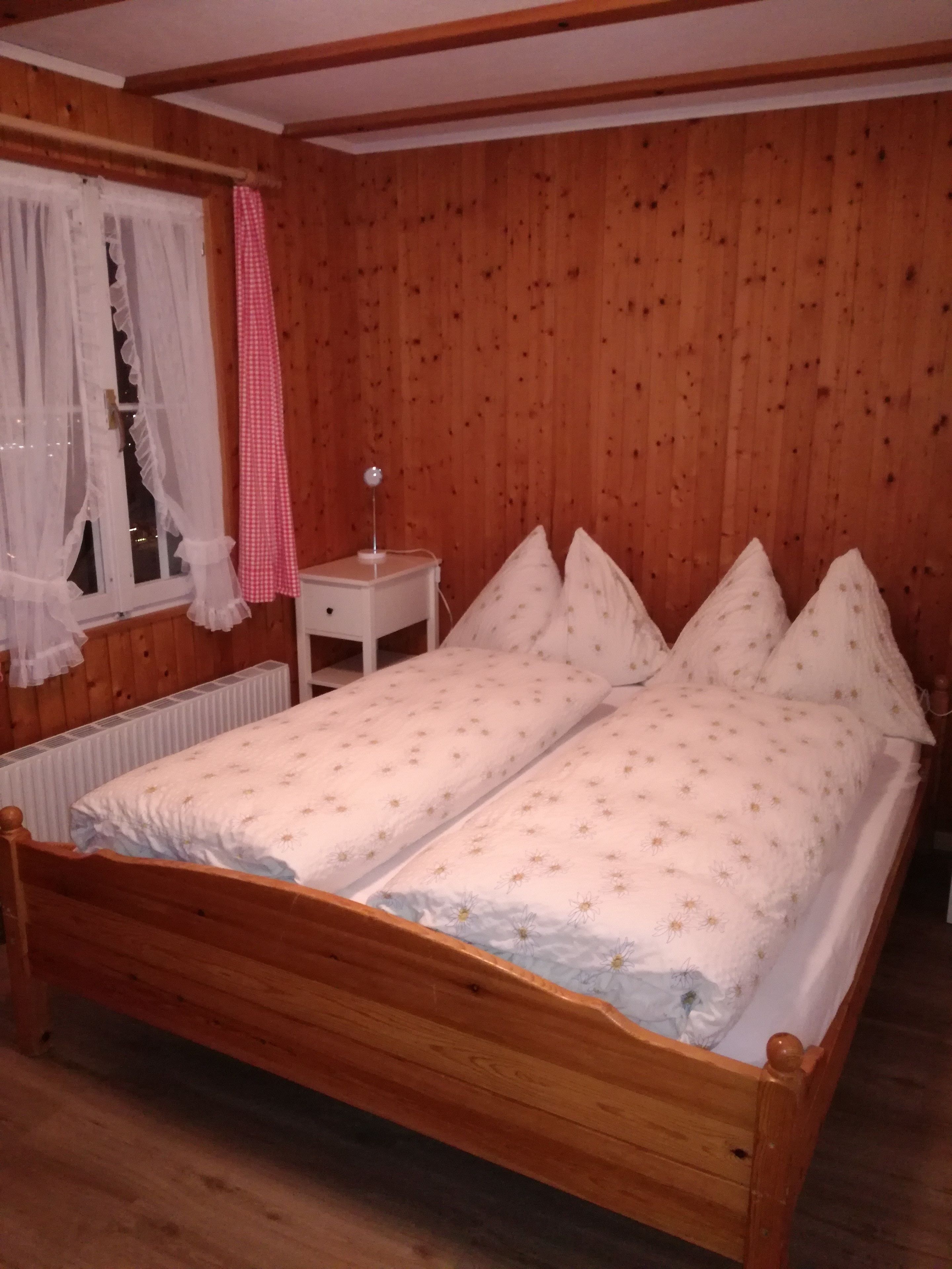 A wooden-paneled room with a double bed, a nightstand, a lamp, and a window with curtains.
