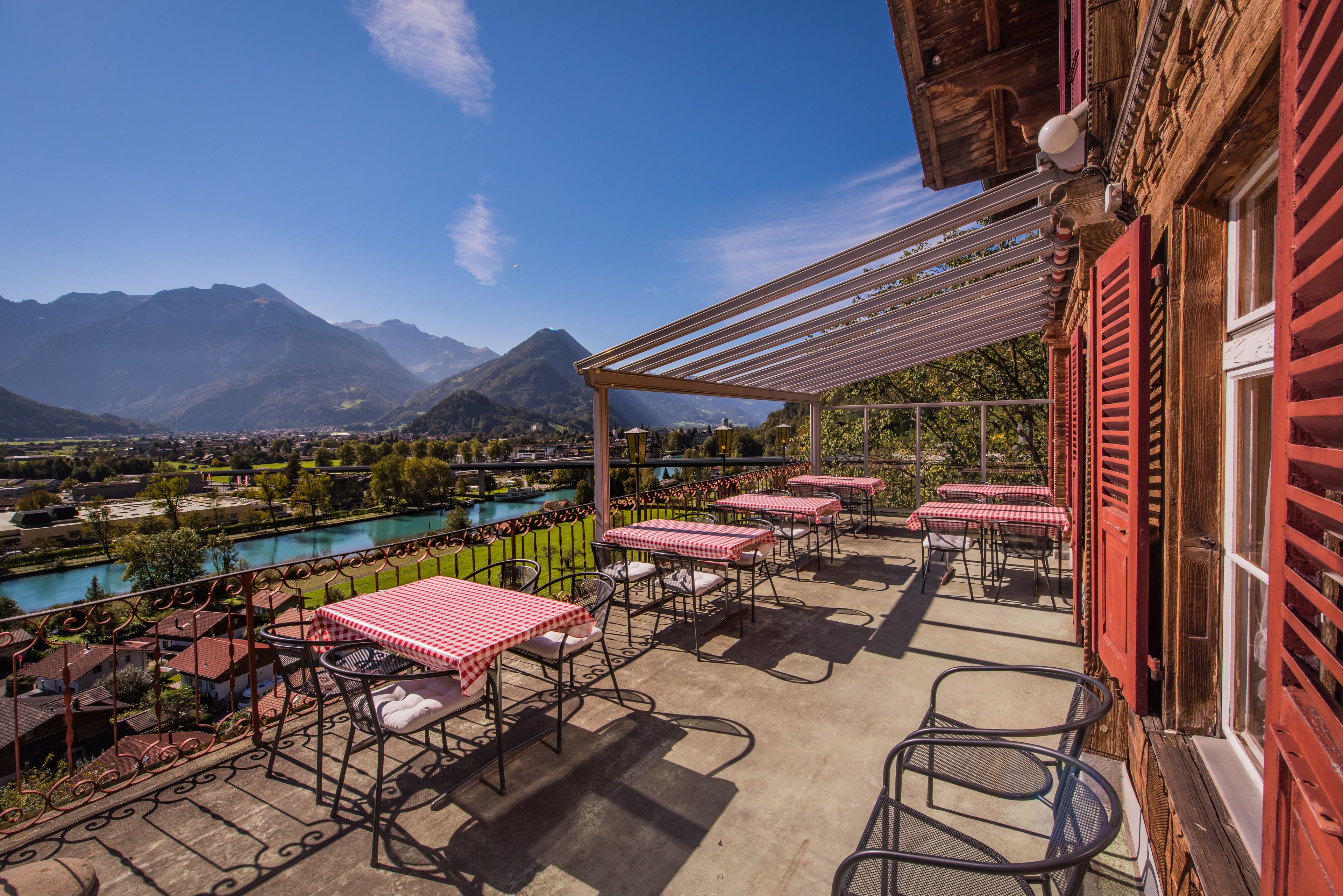 Um terraço com mesas ao ar livre, vista para um rio e montanhas ao fundo.