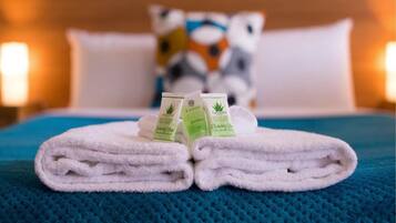 A neatly folded white towel on a blue textured bedspread with two small green bottles labeled "basic" and "cleansing bar" on top.
