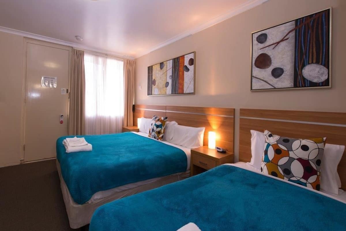 A hotel room with two beds, each with a blue bedspread, white pillows, and a small bedside table with a lamp. There are abstract paintings on the walls and a door with a sign.