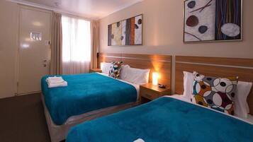A hotel room with two beds, each with a blue bedspread, white pillows, and a small bedside table with a lamp. There are abstract paintings on the walls and a door with a sign.