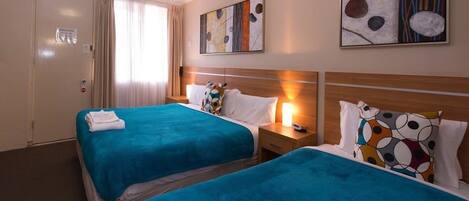 Habitación de hotel con dos camas, cada una con cubrecama azul, almohadas blancas y una mesita de noche con lámpara. Hay pinturas abstractas en las paredes y una puerta con un cartel.