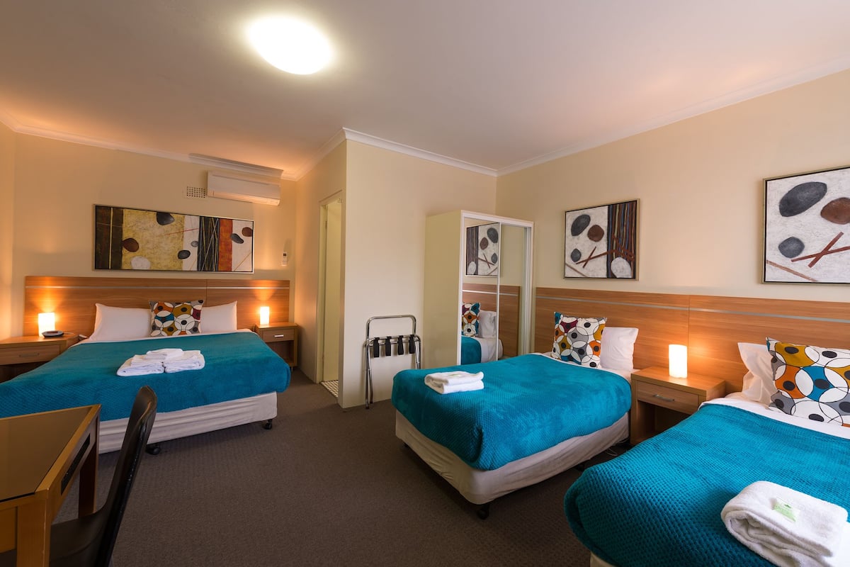 A hotel room with two beds, each with a teal bedspread and white pillows. There are bedside tables with lamps, a mirror, and artwork on the walls.