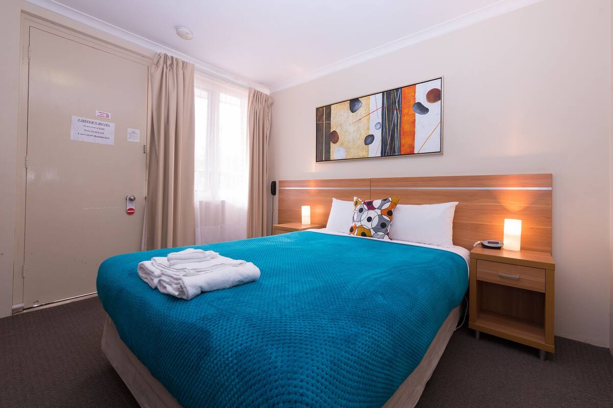 A hotel room with a large bed, a nightstand, a wall-mounted telephone, and a window with curtains.