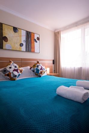 A hotel room with a large bed, a blue blanket, two patterned pillows, a white towel, a bedside table with a lamp, and a painting on the wall.