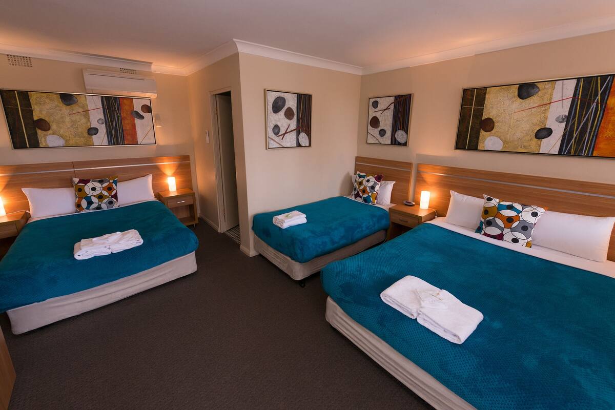 A hotel room with three beds, each with a teal bedspread and white pillows. There are abstract paintings on the walls and an air conditioning unit above the beds.