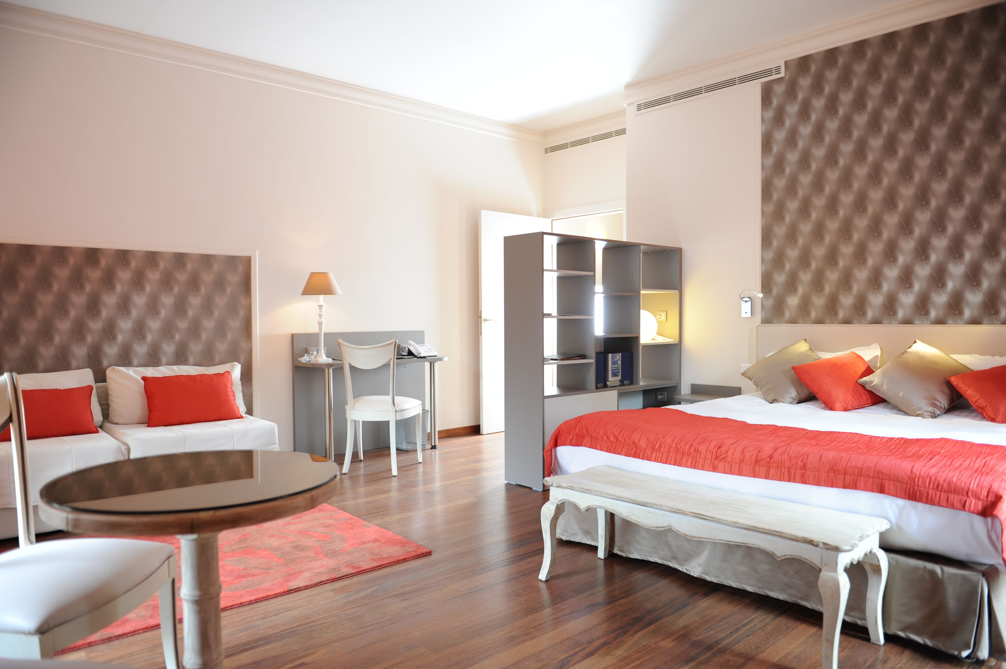 A modern hotel room with a large bed, a seating area with a table, a desk, and a bookshelf.