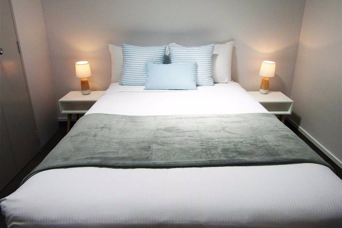 A neatly made bed with a grey blanket, two bedside lamps, and a series of pillows.