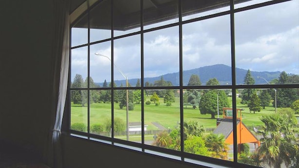 A room with a large window offering a view of a golf course, a mountain, and a building with a sign that reads "MOTEL LOOK". - Pohutu Lodge Motel (Rotorua)