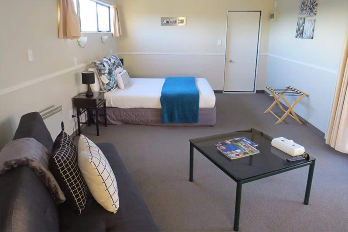 A bedroom with a bed, a bedside table, a nightstand, a window with curtains, a door, a small table with a magazine, a couch, and a wall-mounted drying rack.