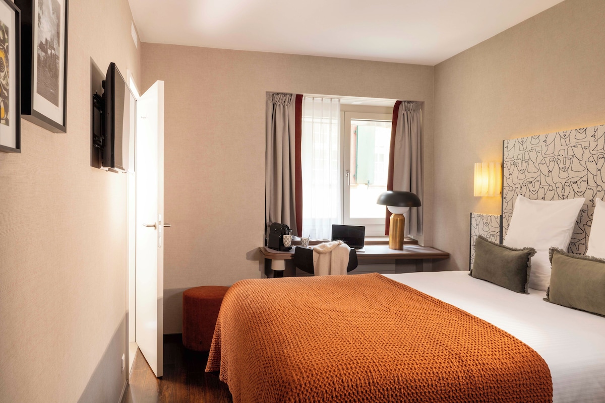 A hotel room with a bed, a desk with a computer, a chair, a window with curtains, and a wall with framed pictures.