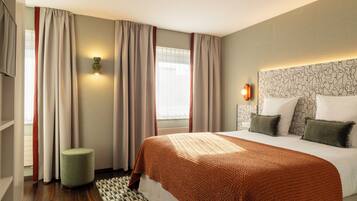 A hotel room with a bed, curtains, a wall-mounted light, a patterned headboard, and a small round ottoman.