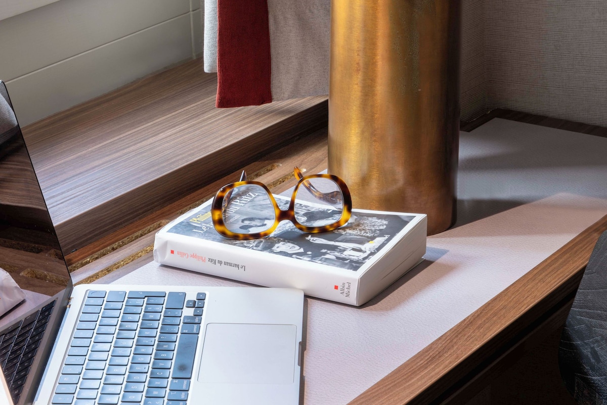 A laptop on a desk with a book and glasses, a lamp, and a window with curtains.