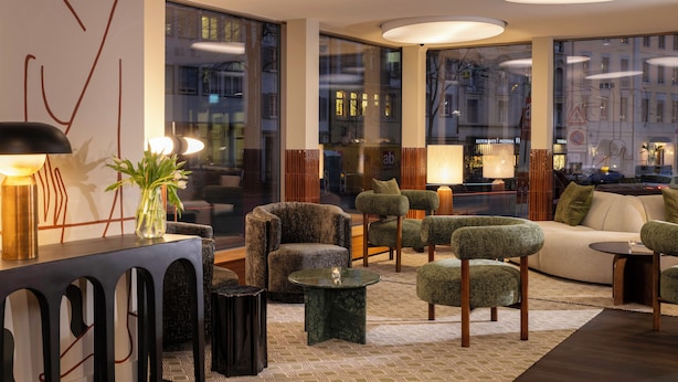 Rebedor modern d'hotel amb diverses zones d'aixovines, incloent un sofà i butaques, una taula de cafè i una consola amb un vas. Grans finestrals ofereixen vistes a la ciutat.