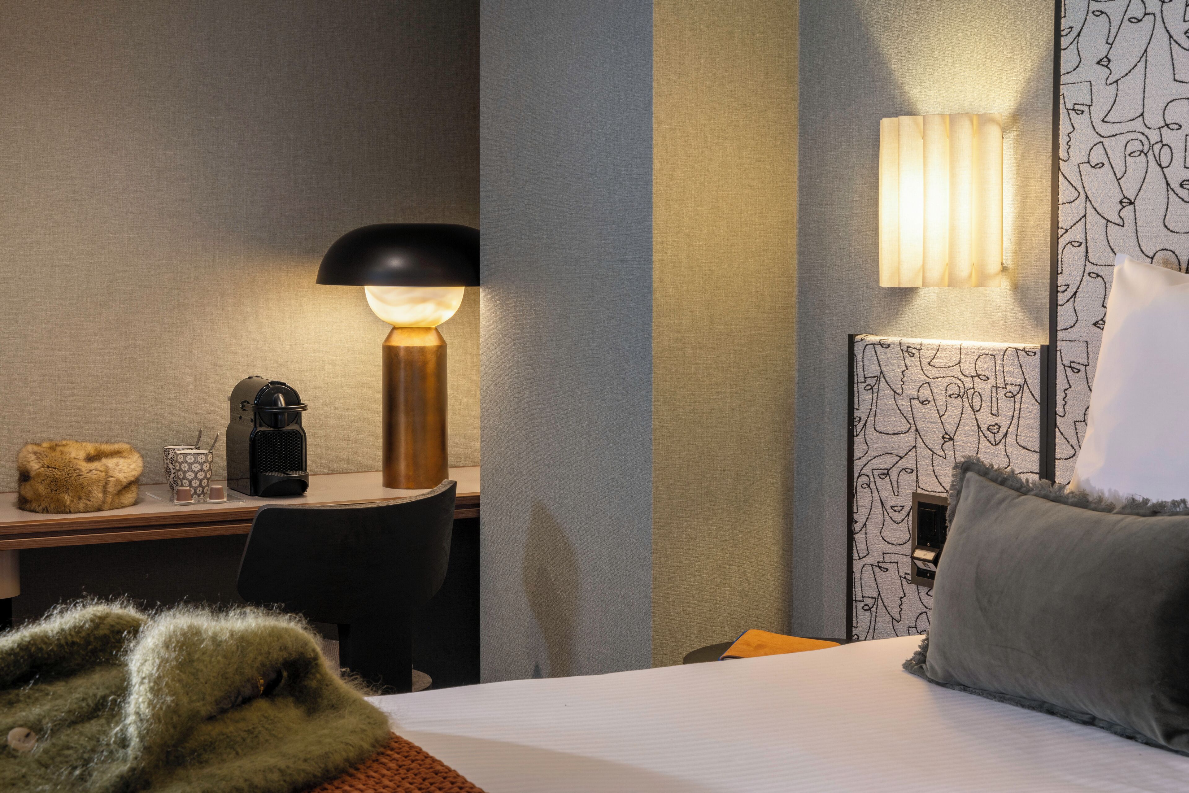 A hotel room with a bed, a desk with a lamp, a coffee maker, and a patterned wall.