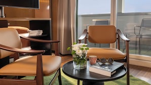 A hotel room with a large window, a flat-screen TV, two leather chairs, a small round table with a book and a vase of flowers, and a bookshelf with books and a lamp. - Hotel D - Basel - Boutique Hotel (Basel)