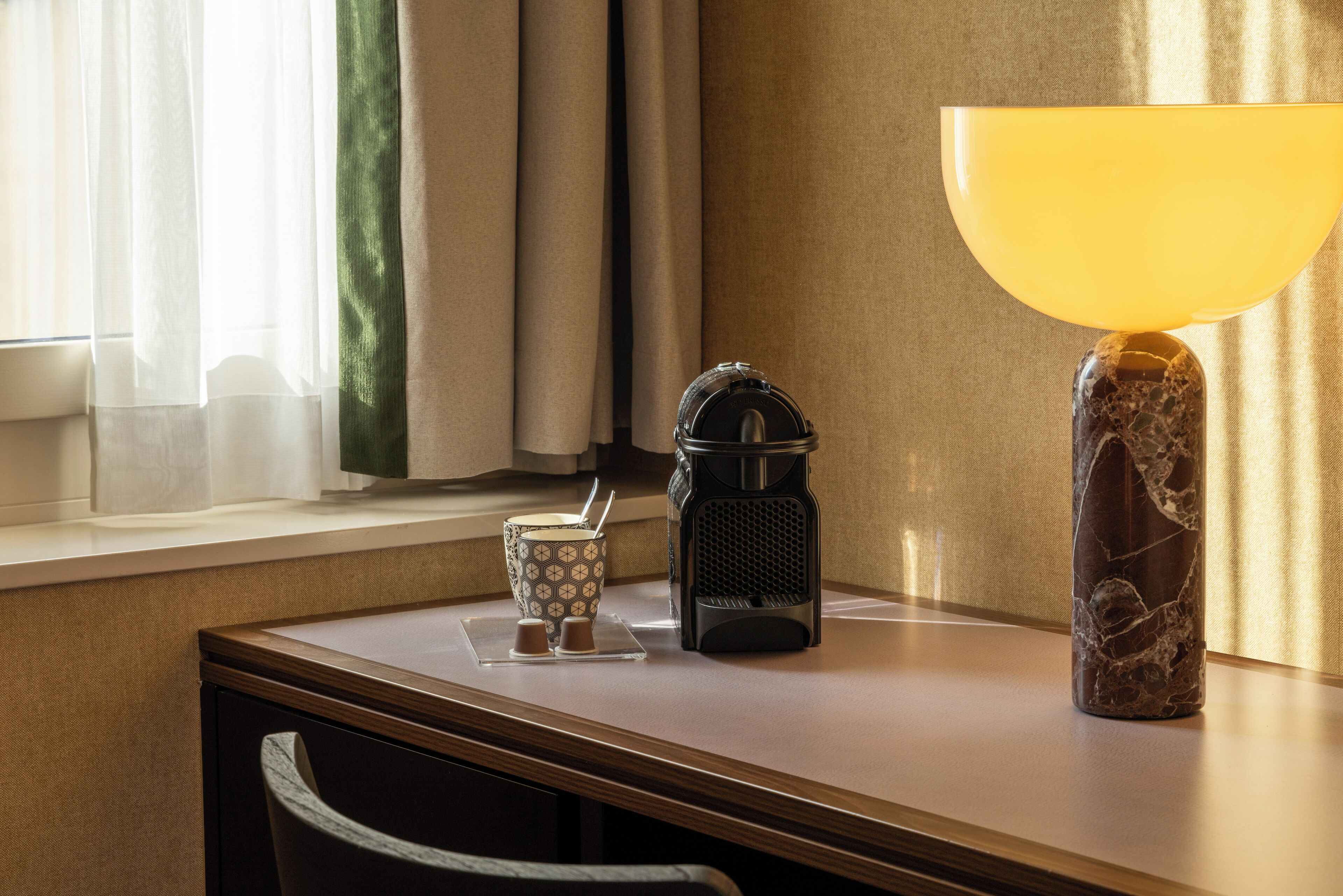 A hotel room with a wooden desk, a coffee maker, a tea set, and a lamp.