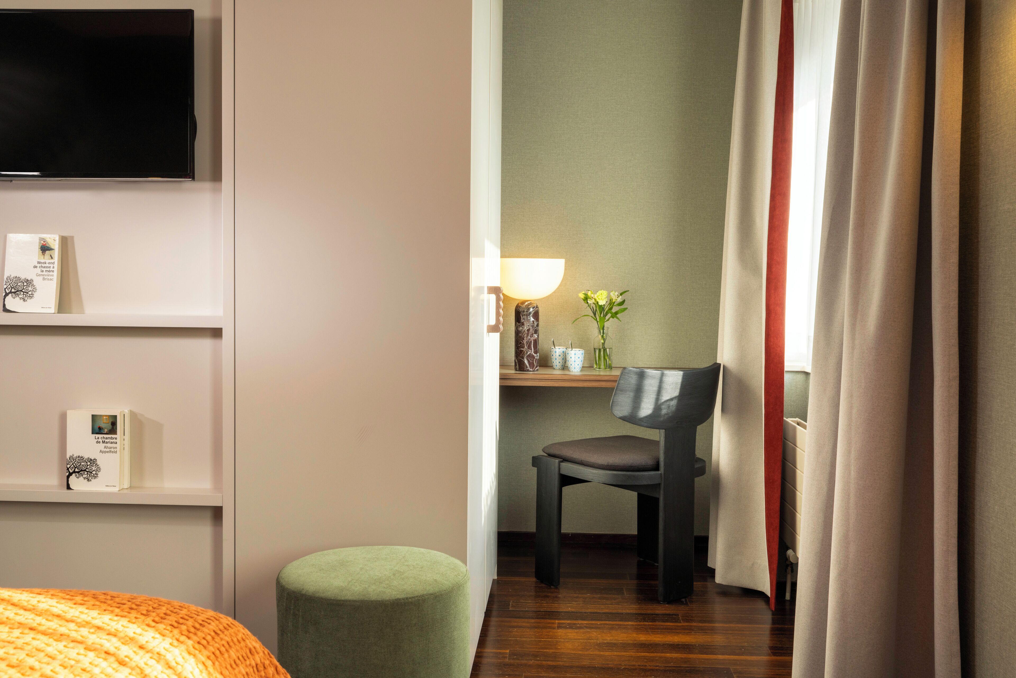 A hotel room with a bed, a desk with a chair, a TV mounted on the wall, and a window with curtains.