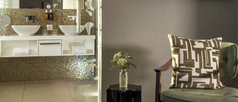 Een moderne badkamer met een glazen douchecabine, een grote spiegel en een wastafel. Er is een flatscreen televisie aan de muur bevestigd en een klein bijzettafeltje met een vaas.