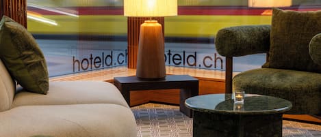 A hotel room with a sofa, armchair, and a small table with a lamp.