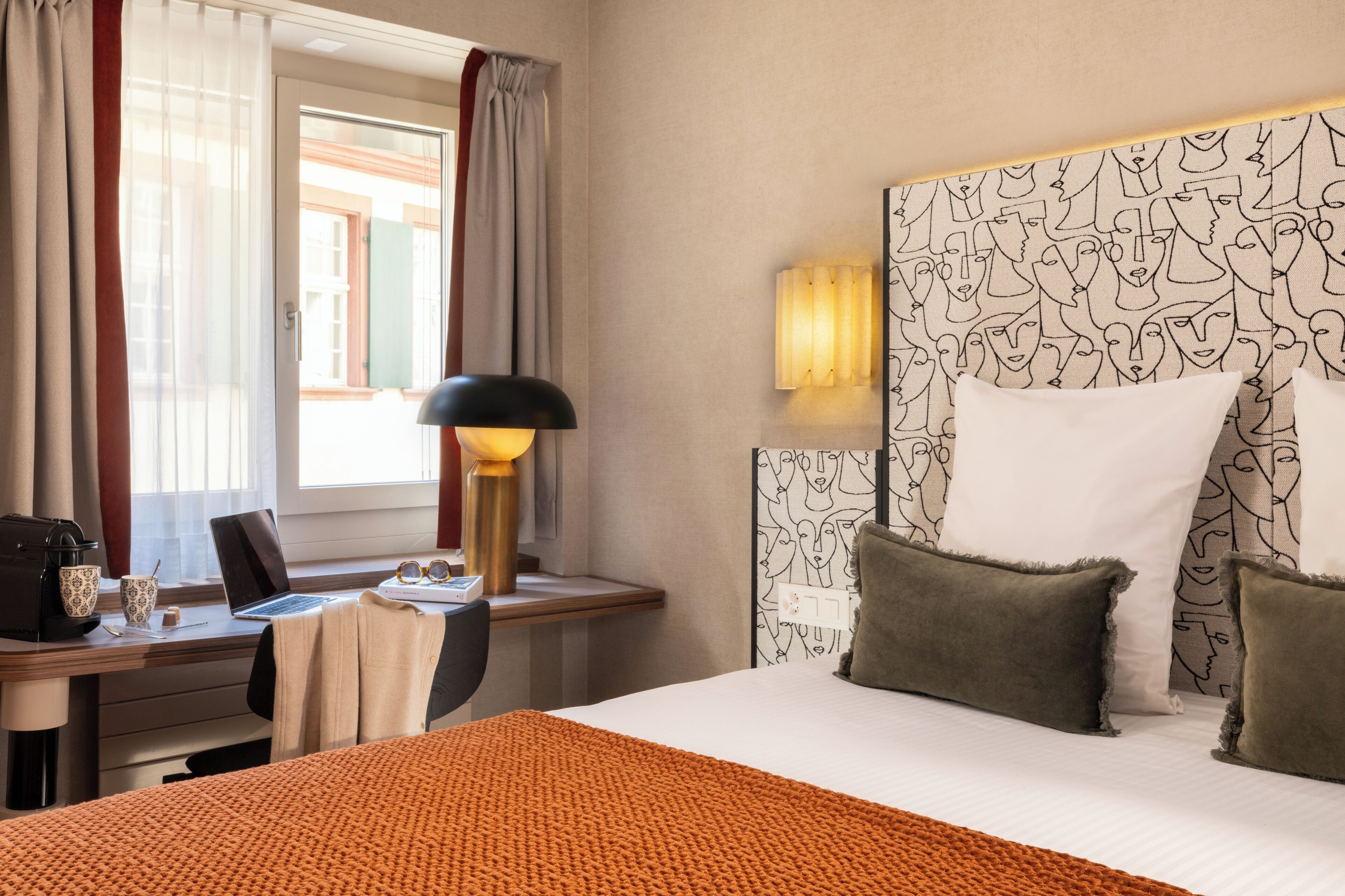 Una habitación de hotel con una cama, un escritorio con una computadora portátil, una lámpara y un ventanal con cortinas.