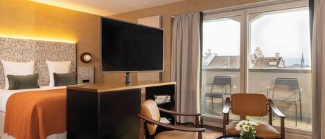 A hotel room with a bed, a desk, a TV, a chair, a table with a vase, and a balcony with chairs.