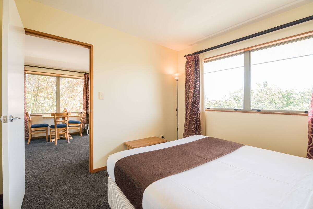 A hotel room with a bed, a desk, and a chair. There is a window with curtains and a view of trees outside.