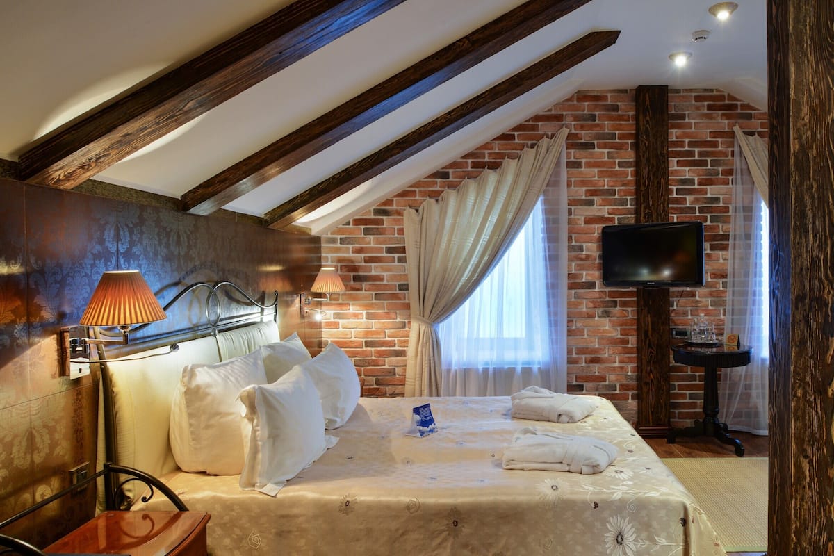 A cozy attic bedroom with exposed wooden beams, a brick wall, a bed with white linens, a TV, and a small table.