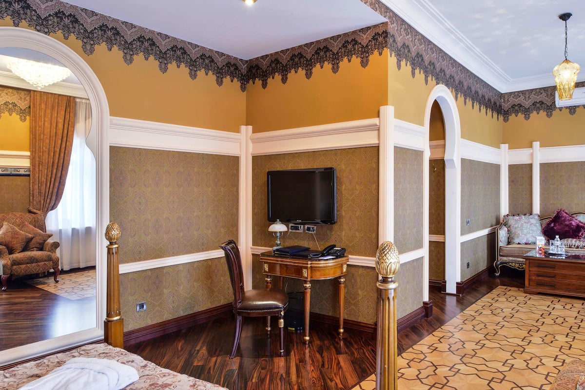 A room with a large mirror, a wooden chair, a sofa, a small table with a lamp, and a television.