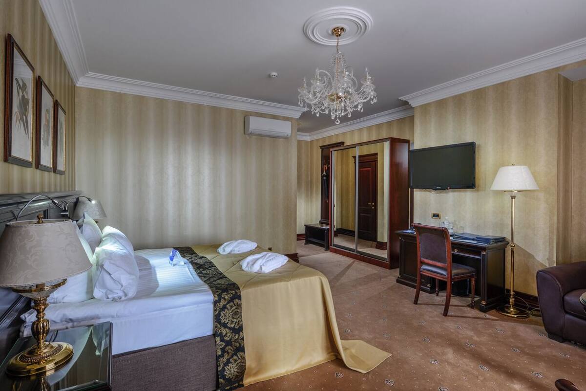 A hotel room with a bed, a desk, a chair, a TV, a lamp, and a chandelier.