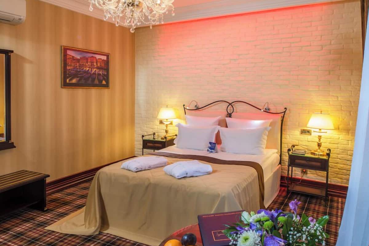 A hotel room with a bed, bedside lamps, a chandelier, a plaid-patterned carpet, a round table with fruit and flowers, and a framed picture on the wall.