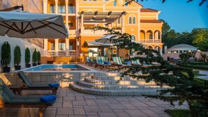 Uma área de piscina com espreguiçadeiras e guarda-sóis, cercada por um edifício com várias varandas e uma fonte.
