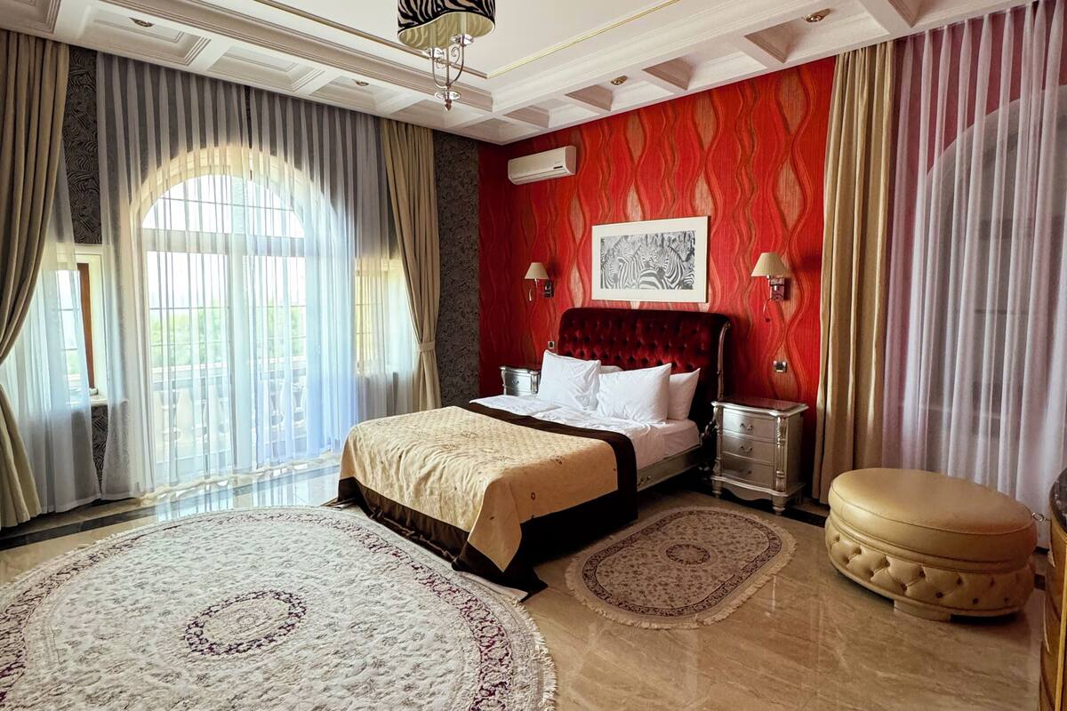 A spacious bedroom with a large bed, a sitting area with a cushioned ottoman, and a large window with curtains.