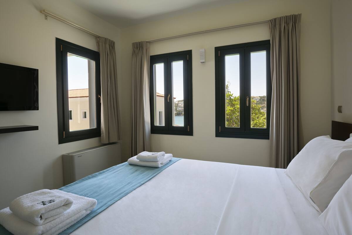 Une chambre d’hôtel avec un lit, deux fenêtres avec des rideaux, un téléviseur à écran plat fixé au mur et une vue sur l’extérieur.