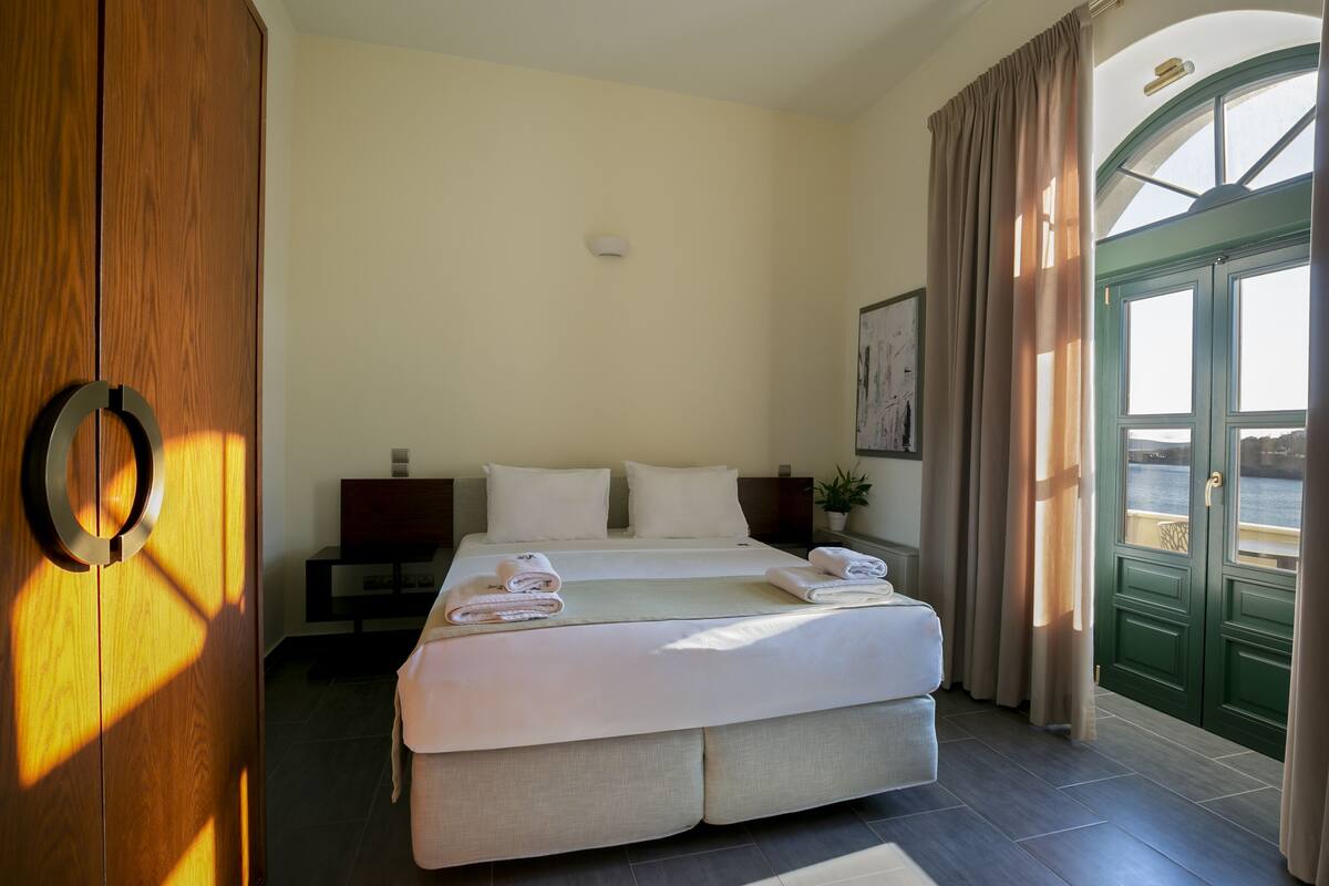 Une chambre d’hôtel avec un grand lit, une armoire en bois, une table de chevet et une vue sur la mer, visible à travers une porte verte.