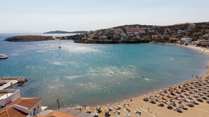 Un pueblo costero con una playa bordeada por tumbonas y sombrillas, un barco anclado cerca de la orilla, y edificios en una colina.