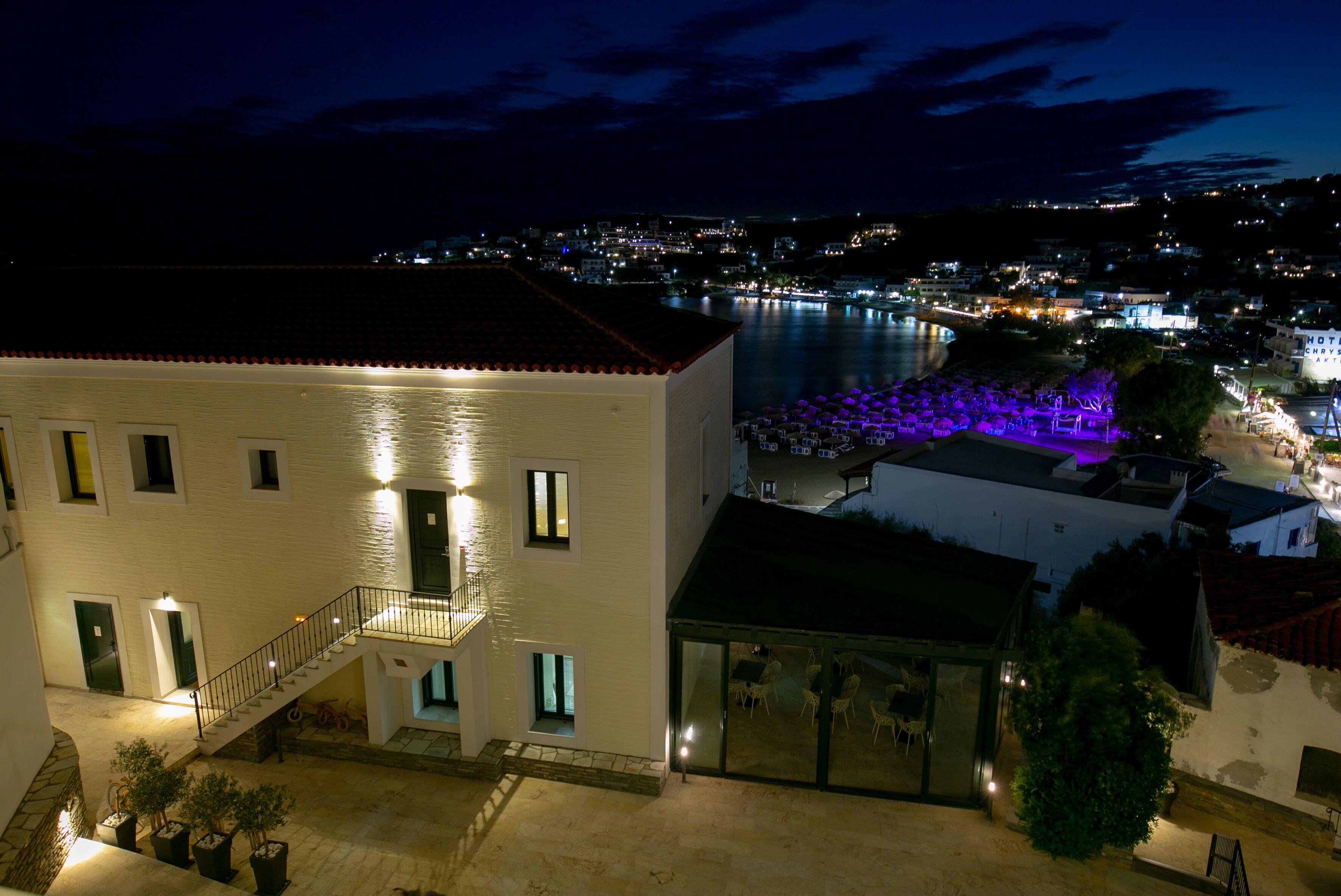 A hotel with a balcony overlooking a body of water, a pool area with umbrellas, and a staircase leading to the upper floor.