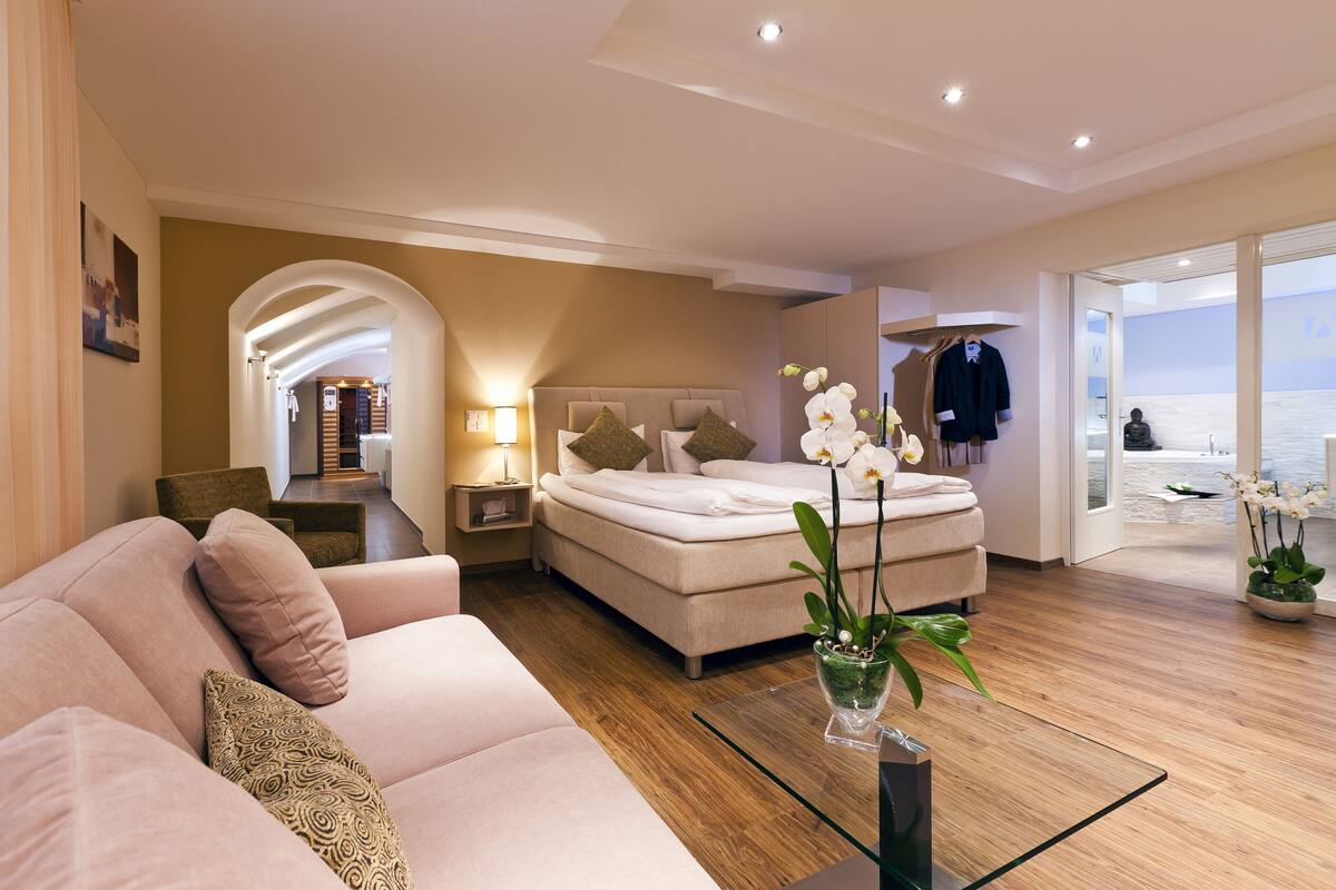 A modern hotel room with a bed, a sofa, a glass coffee table, and a view of the beach.