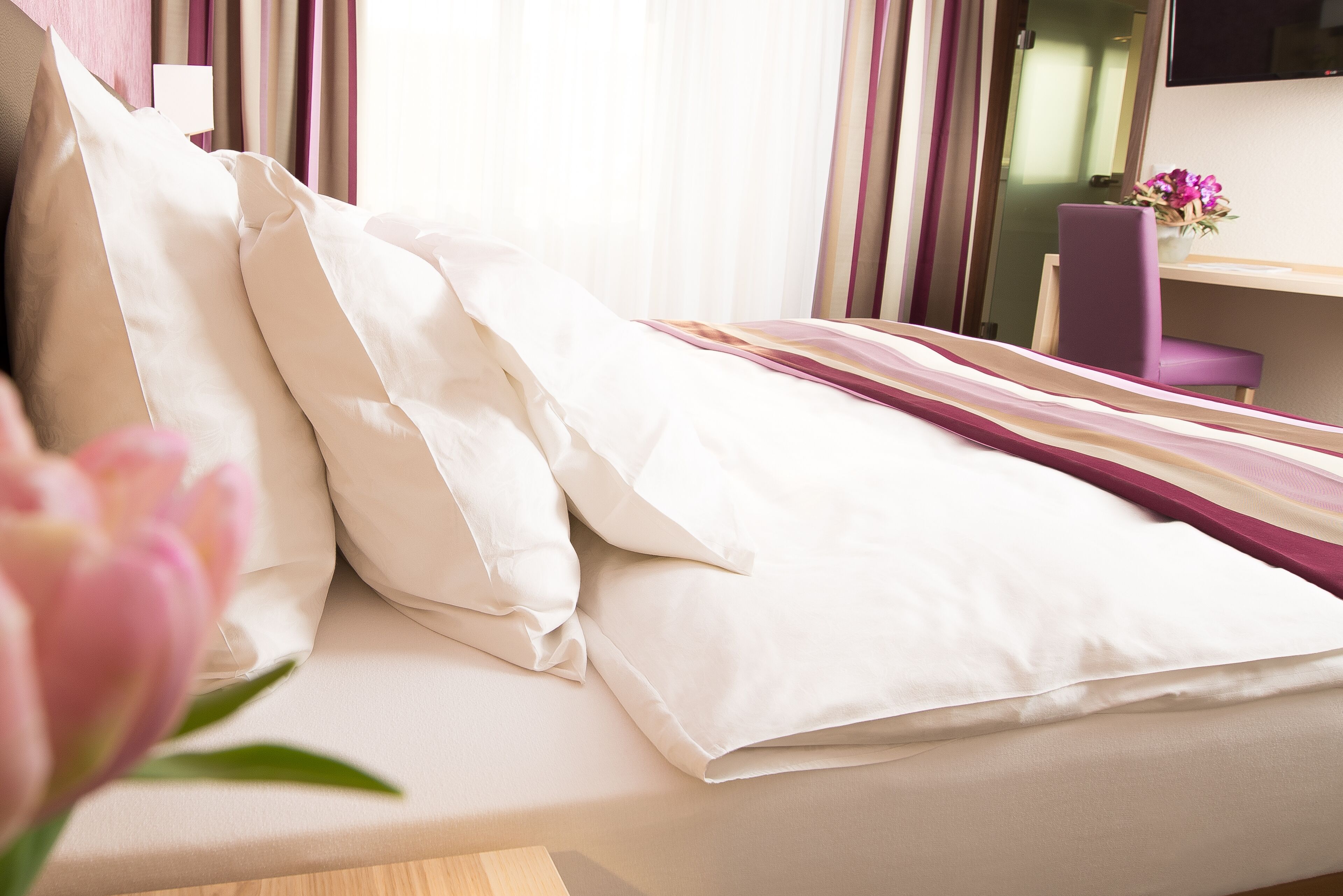 A neatly made bed with white linens, a purple chair, and a small table with a vase of flowers.