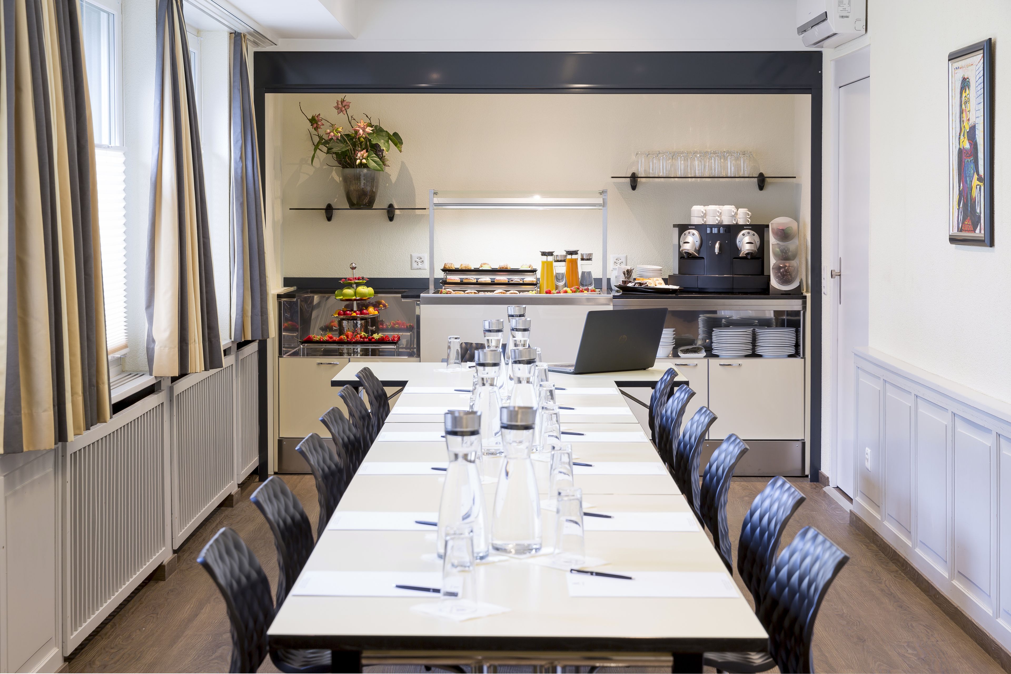 Une salle de conférence équipée d’une longue table, de chaises, d’une machine à café et d’un coin cuisine avec des étagères et des appareils électroménagers.