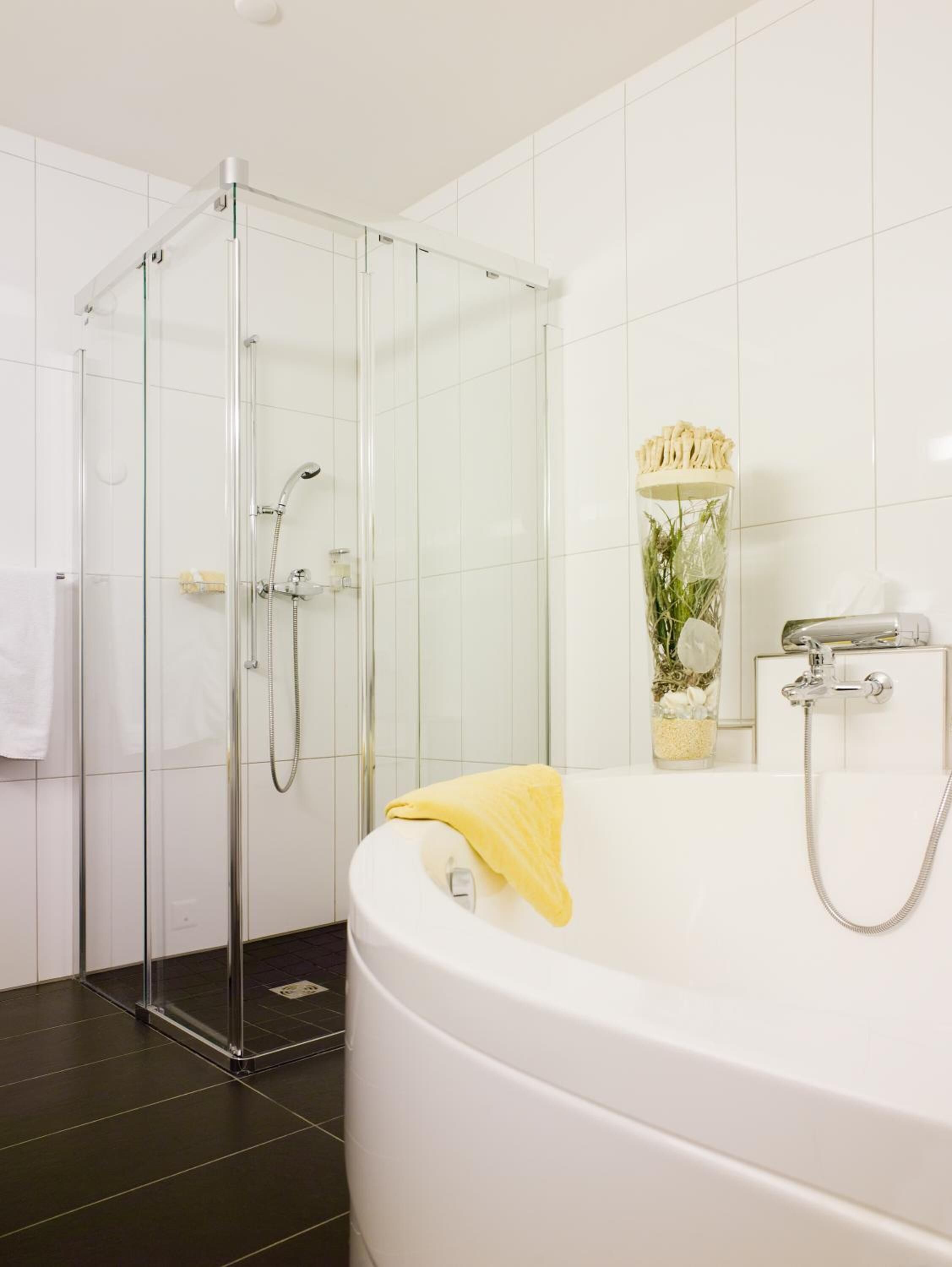 Un baño moderno con cabina de ducha de vidrio, una bañera blanca y una toalla amarilla, además de un florero con flores en la pared.