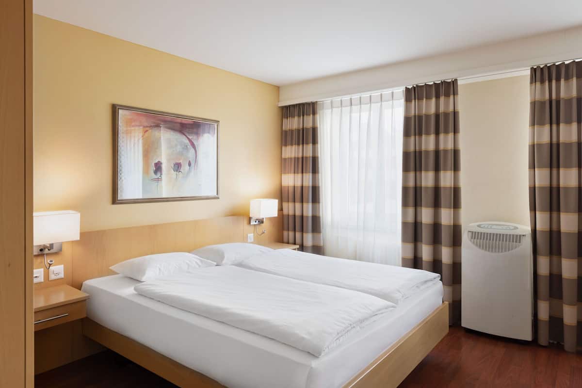 A hotel room with a large bed, two bedside tables with lamps, a wall-mounted artwork, a window with curtains, and a standing heater.