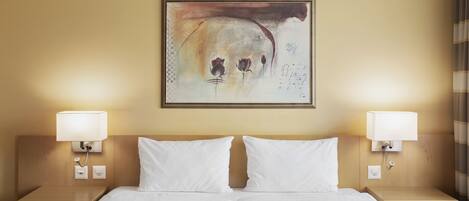 A hotel room with a double bed, two bedside tables, a wall-mounted abstract painting, and two wall-mounted lamps.
