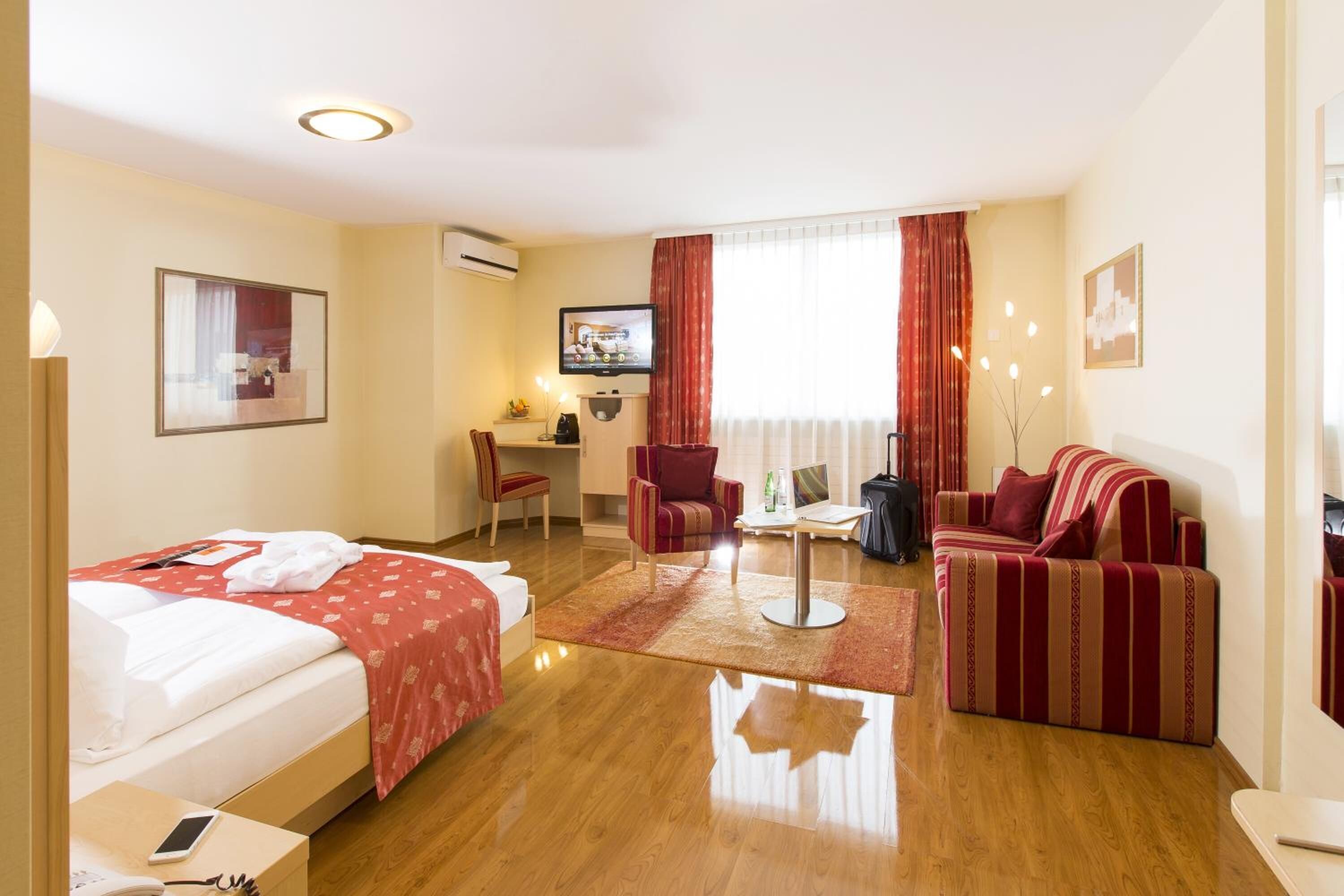 A hotel room with a bed, a sofa, a desk with a chair, a TV, and a window with curtains.