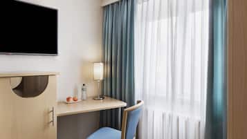 A hotel room with a flat-screen TV mounted on the wall, a desk with a lamp, a chair, and a window with sheer curtains.