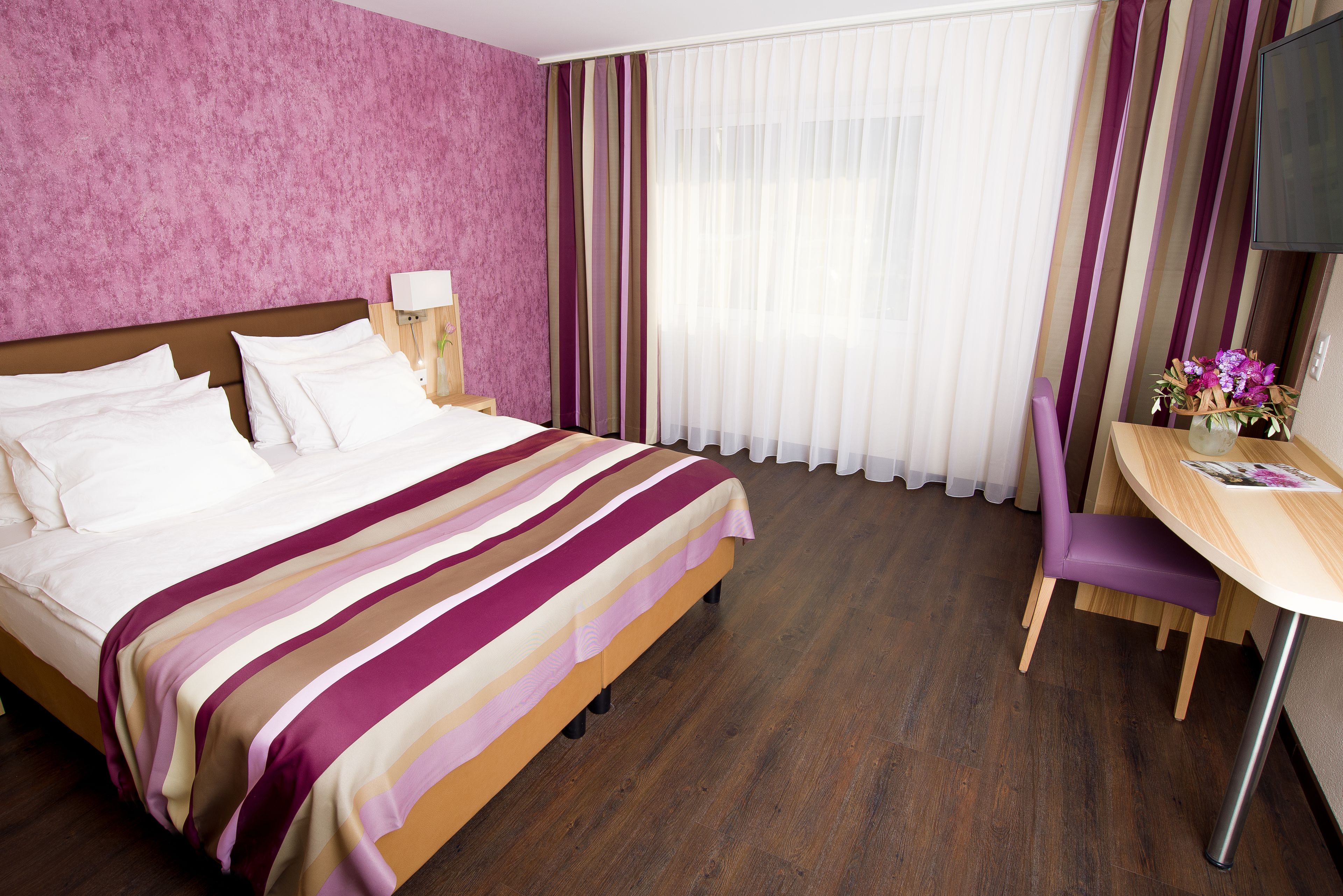 A hotel room with a large bed, a desk with a chair, a TV, and a window with curtains.