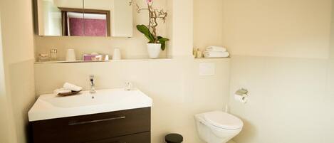 Un baño moderno con lavamanos blanco, mueble oscuro, espejo, inodoro y un cubo de basura.