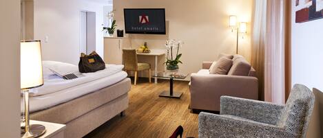 Una habitación de hotel con cama, sofá, sillón, escritorio y televisor mostrando el nombre del hotel.